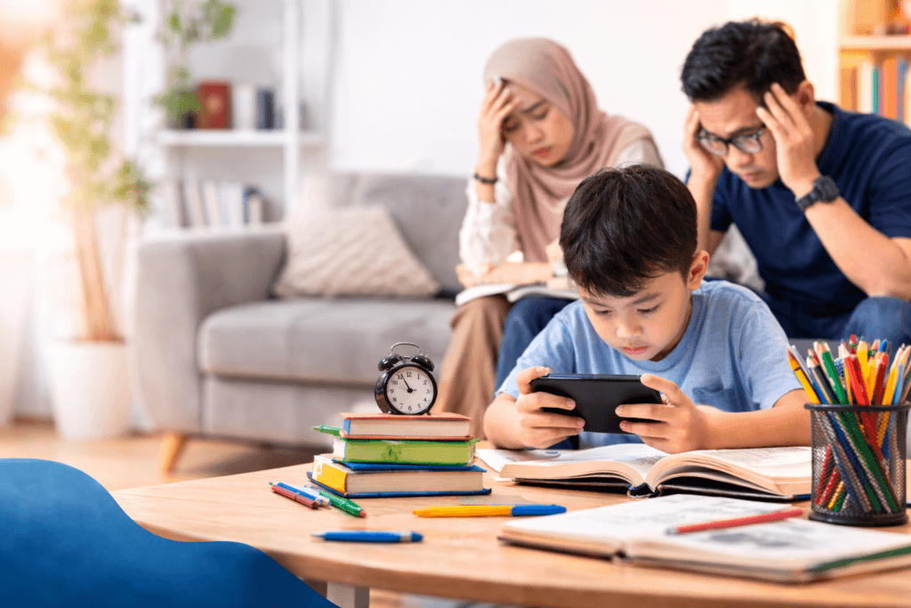 Anak bermain gadget di meja belajar sementara orang tua terlihat khawatir karena dampak kecanduan gadget pada anak
