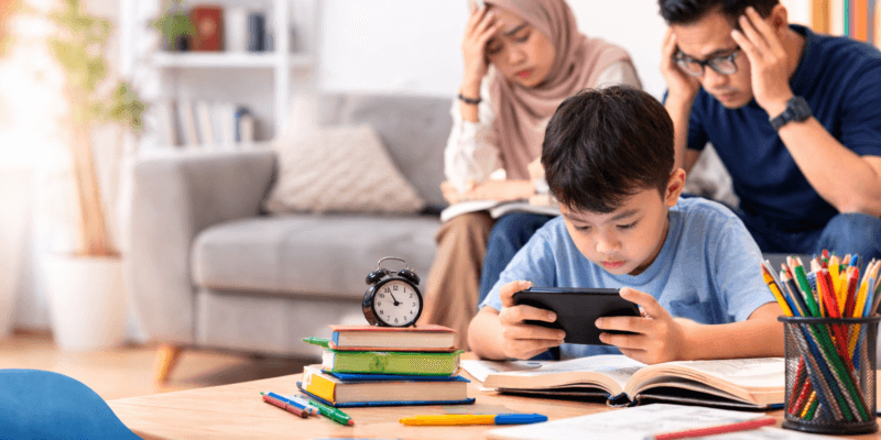 Anak bermain gadget di meja belajar sementara orang tua terlihat khawatir karena dampak kecanduan gadget pada anak