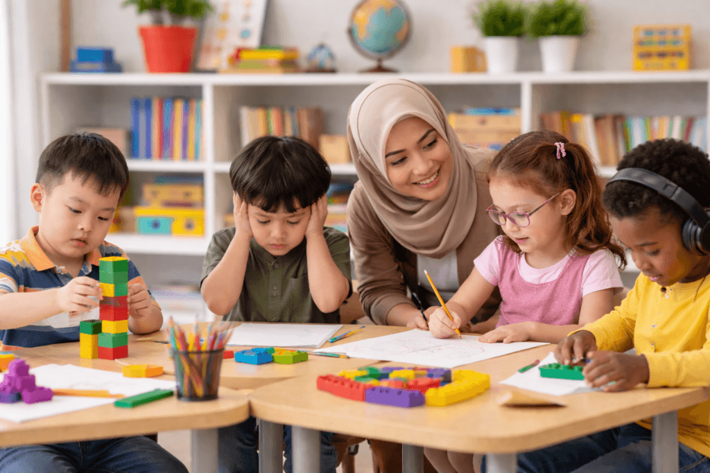 Guru wanita Indonesia berhijab membantu beberapa anak berkebutuhan khusus belajar bersama di ruang kelas dengan mainan edukatif dan buku di meja.