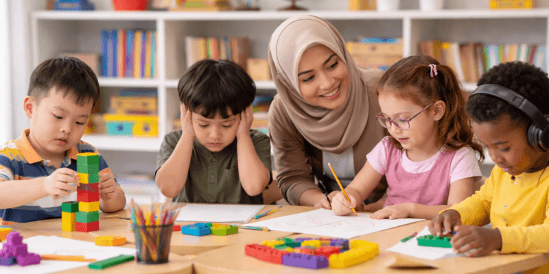Guru wanita Indonesia berhijab membantu beberapa anak berkebutuhan khusus belajar bersama di ruang kelas dengan mainan edukatif dan buku di meja.