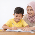 Guru wanita Indonesia berhijab membantu siswa belajar menulis di meja belajar dalam suasana homeschooling yang nyaman di rumah.