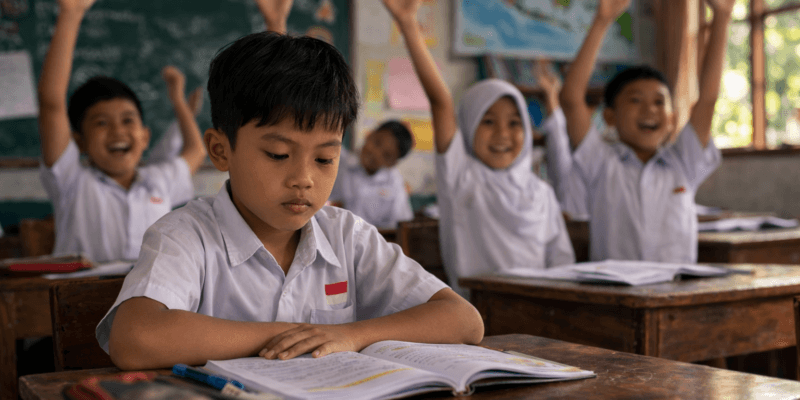 Seorang siswa berwajah Jawa duduk diam di dalam kelas sekolah dengan suasana belajar yang tenang, terlihat fokus namun pendiam saat mengikuti pelajaran di ruang kelas bersama guru.
